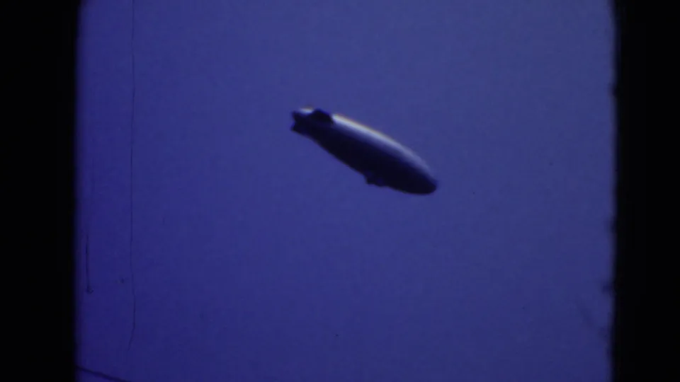 A blimp is framed low against the blue sky, with dark film borders and soft 8mm texture visible.
