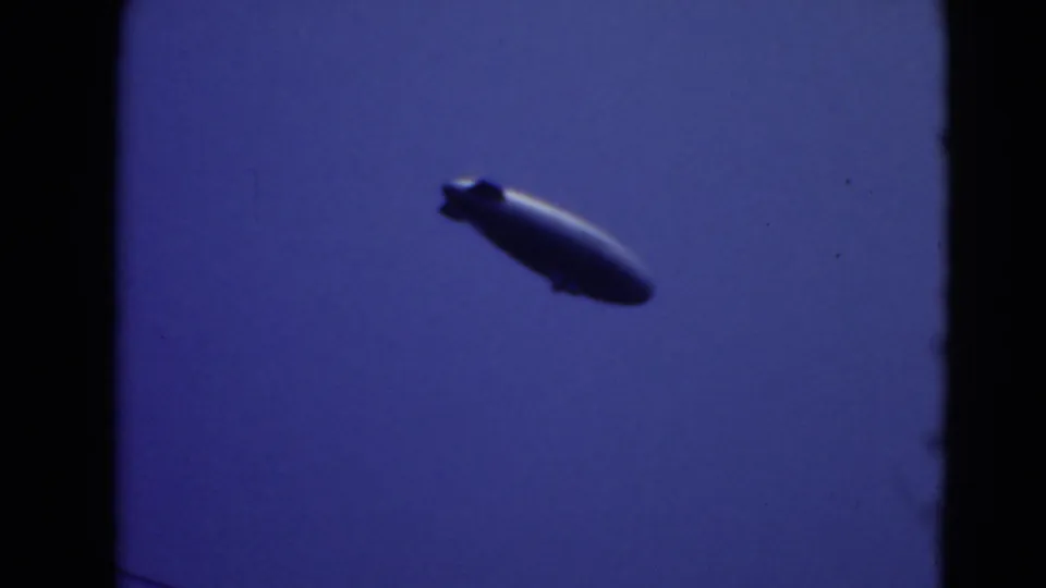Final cataloged frame shows the blimp farther across the sky in soft, blue-toned 8mm footage.