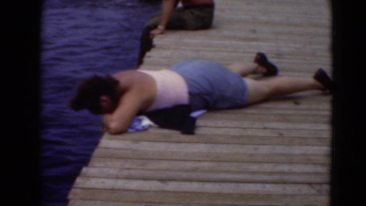 Adult in a light tank top and shorts lies on a wooden dock beside blue water.