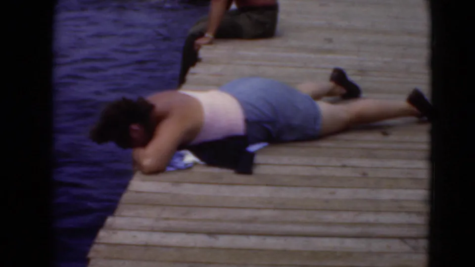 In archival home-movie footage from Middletown, Pennsylvania, an adult lies on a dock near blue water in summer clothing.