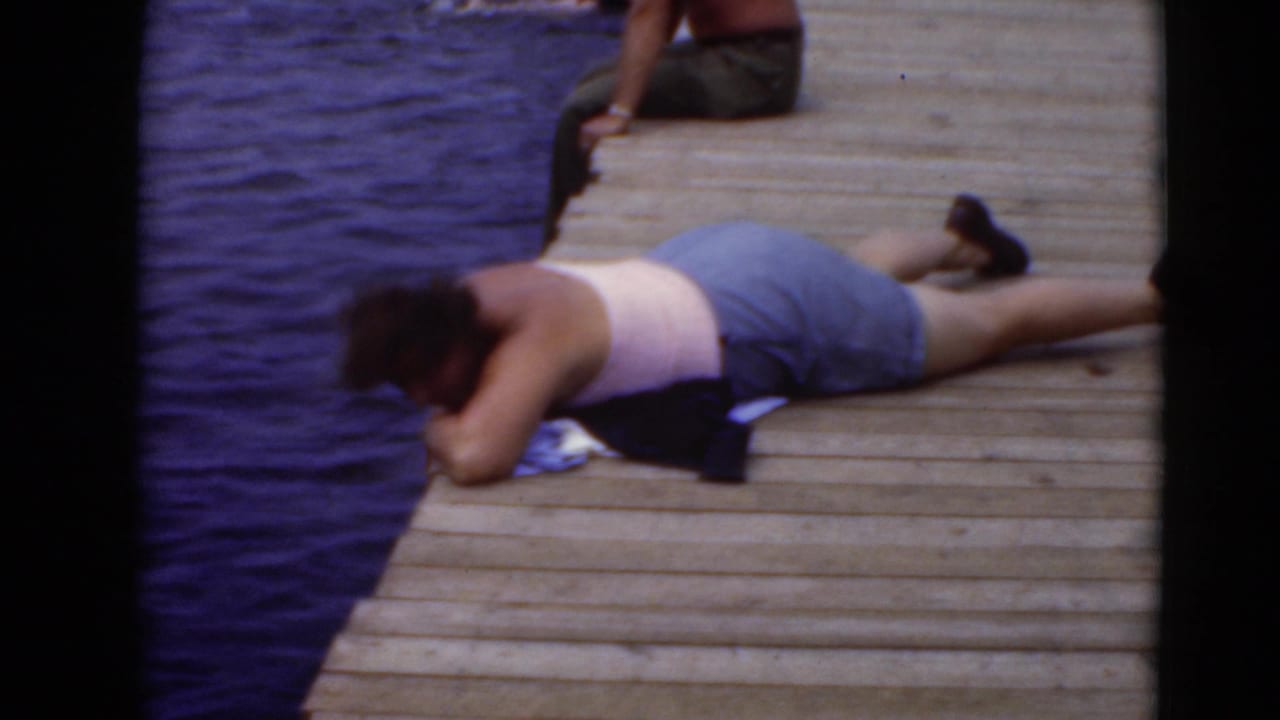 A person in a light top and shorts lies on a wooden dock beside calm water in the 8mm home-movie frame.