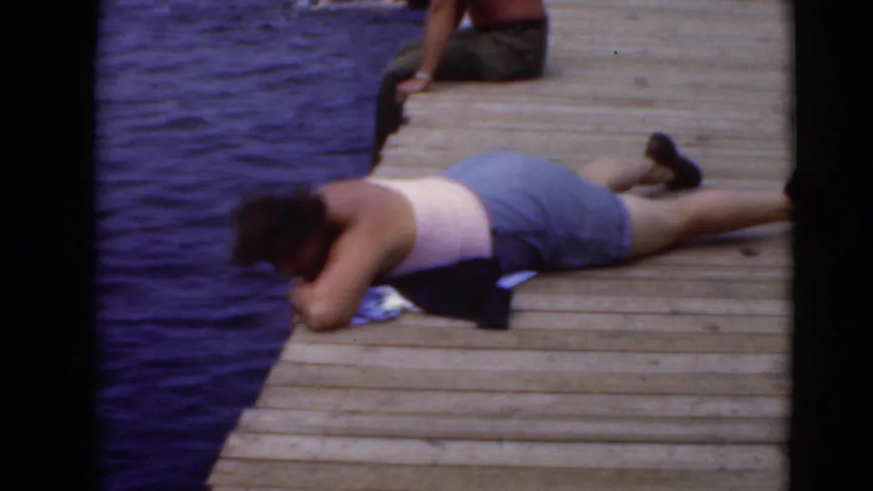 A person in a light top and shorts lies on a wooden dock beside calm water in the 8mm home-movie frame.