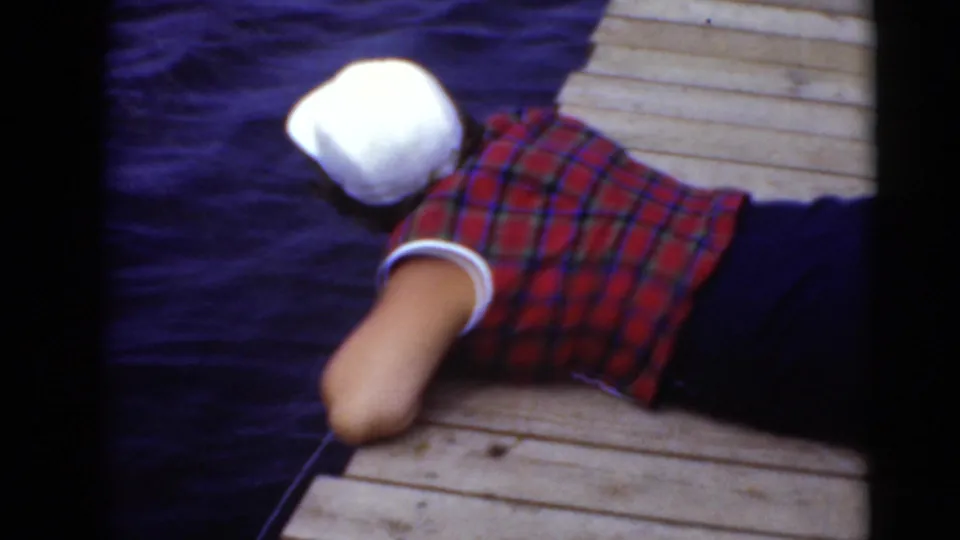 A nearby adult in a red plaid shirt and white cap lies on the wooden dock near the water in the home-movie scene.