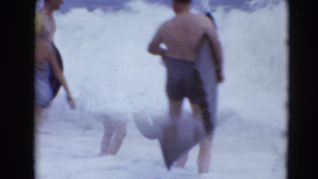 In supplied 1947 home-movie footage, two beachgoers stand in breaking surf as one carries a dark inflatable object.