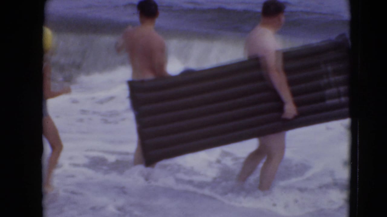 Two shirtless men stand in shallow surf, one holding a long dark inflatable mattress.