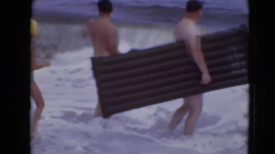 Two men in swim trunks wade near shore, with one carrying a long dark inflatable mattress across the frame.