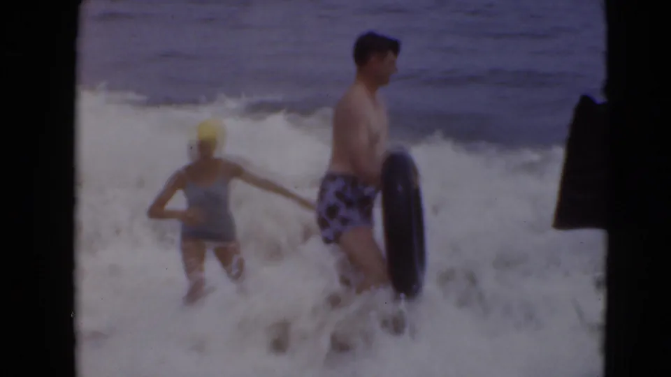 A person wearing a yellow swim cap wades beside a man holding an inner tube as surf breaks around them.