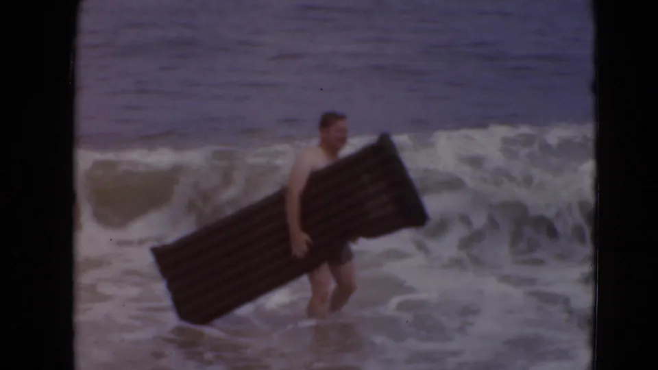 A beachgoer in swim trunks holds a long dark inflatable mattress while standing in foamy water near shore.