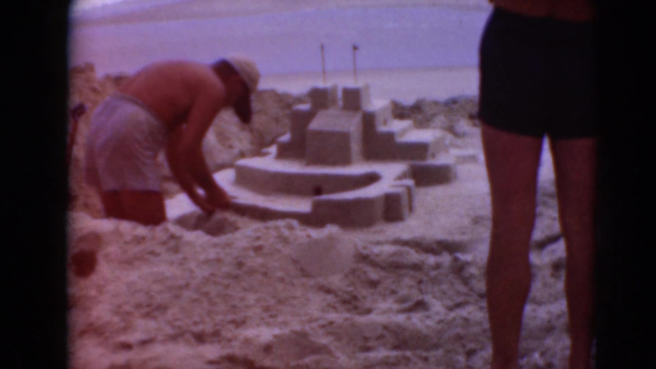 In archival 8mm footage, a man works on a detailed sandcastle as a woman in a dark swimsuit stands near the beach scene.