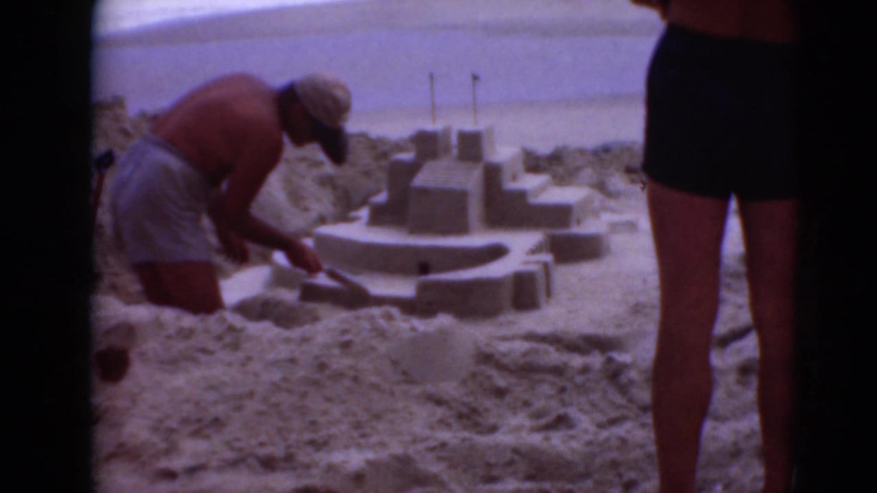 Man in swim trunks shapes a sandcastle while a standing figure is partly visible beside him.