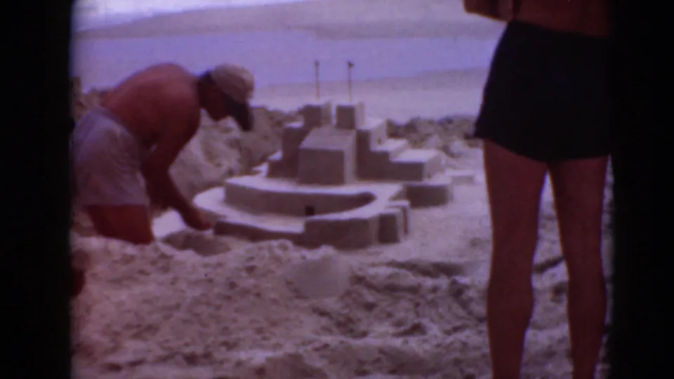 The home-movie frame shows a man tending a sandcastle in the foreground while a woman in a dark swimsuit stands between camera and beach scene.