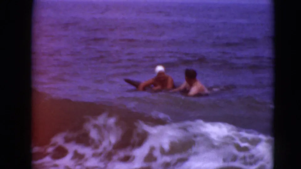 Two adult swimmers are in the surf, one wearing a white cap and holding or lying near a board as waves break in the foreground.