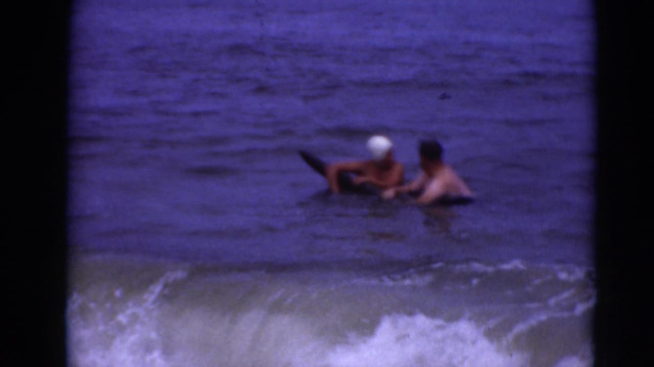 Two people in shallow surf hold onto a dark board as waves roll behind them.