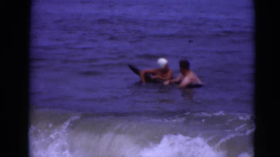 Two people, one wearing a white swim cap, steady themselves on a dark board or float in shallow surf in archival 8mm footage.