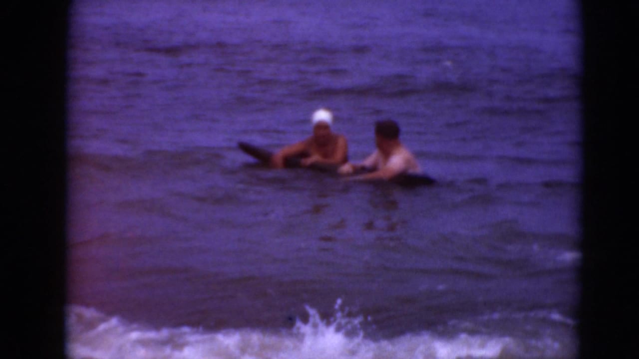 Two people remain in the water with a dark float as surf gathers in the foreground.