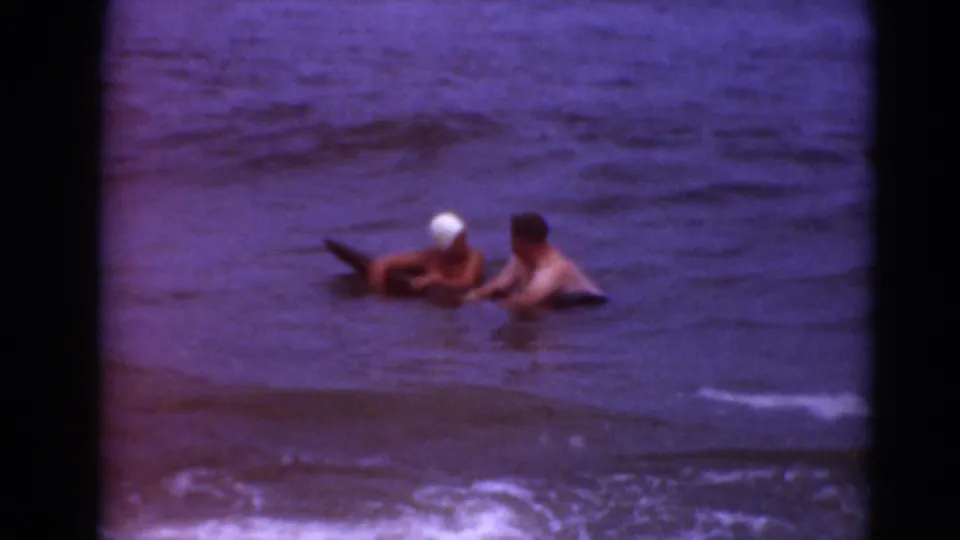Two people appear seated or crouched around a dark board or float in shallow surf, framed by the soft grain of 8mm film.