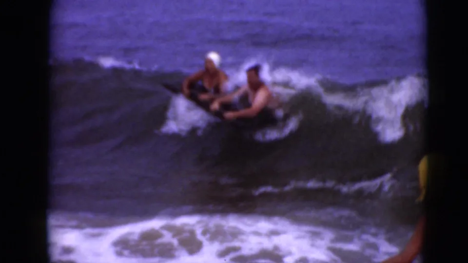 Two people on or beside a dark board meet a breaking wave, with white foam surrounding them in a soft 8mm frame.