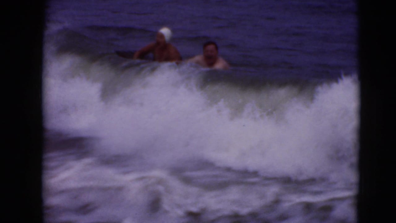 1947 8mm home-movie frame of two swimmers playing in rough surf, one in a white swim cap, with waves breaking around them.