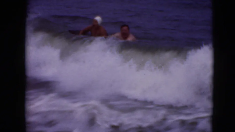 1947 8mm home-movie frame of two swimmers playing in rough surf, one in a white swim cap, with waves breaking around them.