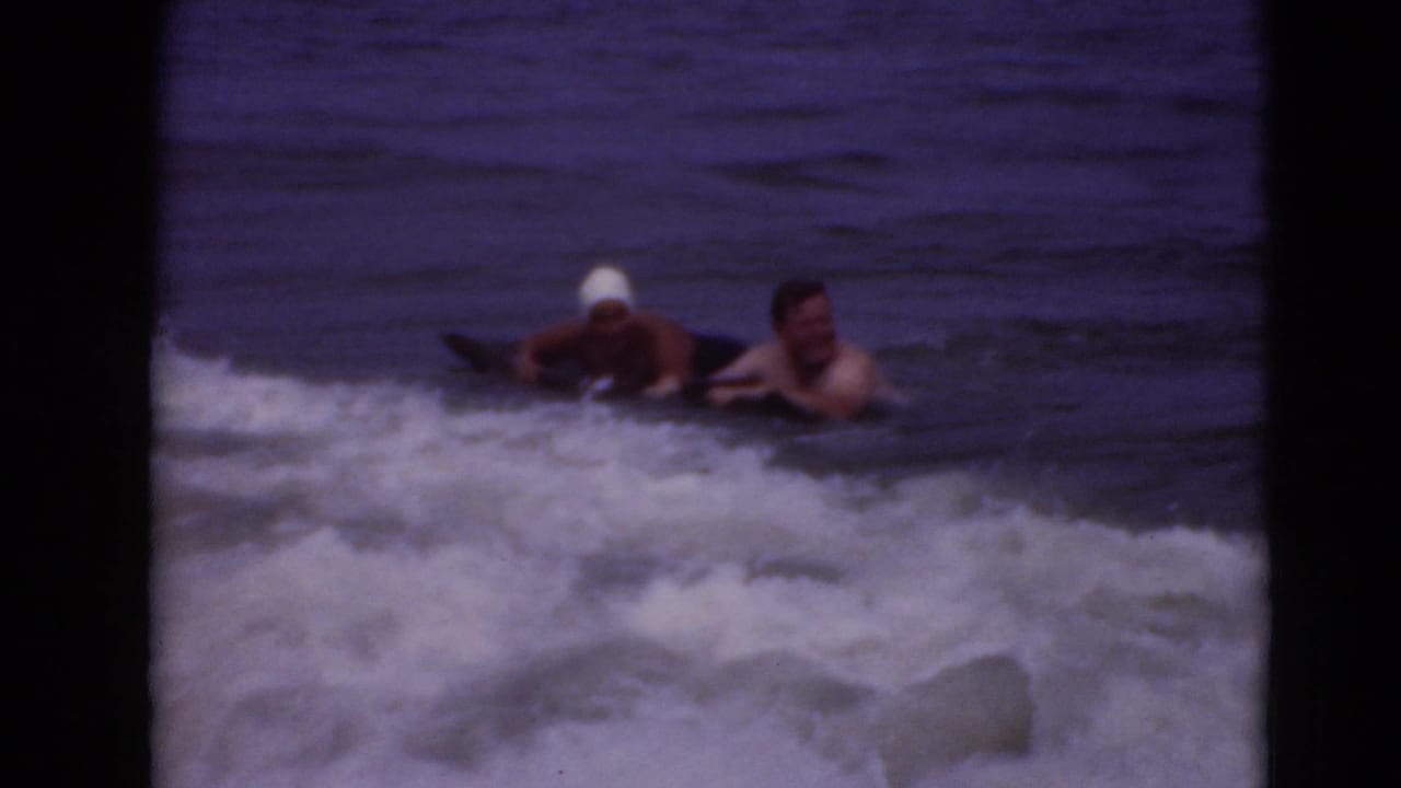 Two swimmers lie near a dark board in choppy water as a wave rolls behind them.