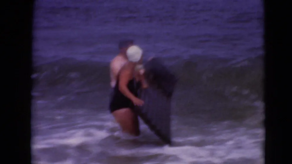 1947 8mm home-movie frame of a swimmer in a white cap standing in shallow waves beside a dark board.