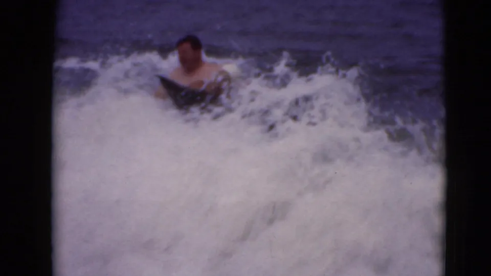 1947 8mm frame of a swimmer with a dark board amid breaking white water, filmed from a shore-side viewpoint.