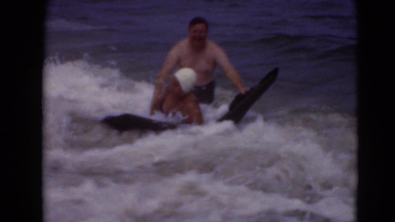 Two adults in rough surf beside a black bodyboard, one wearing a white bathing cap.