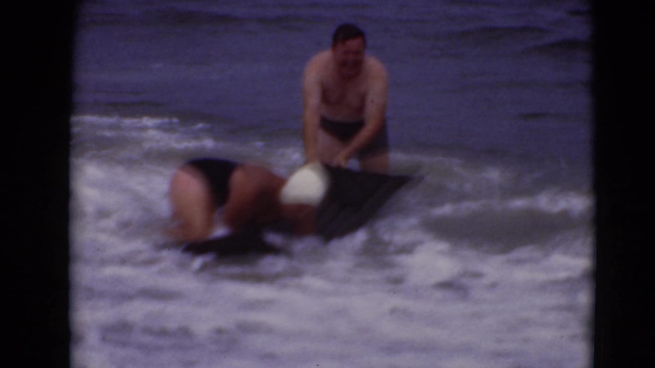 A man stands in the surf while another person bends over a black bodyboard in breaking waves.