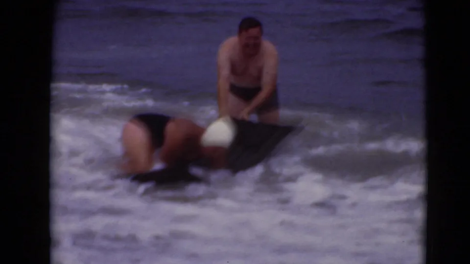 1940s 8mm frame showing an adult man in dark trunks and another adult low over a black bodyboard amid churning surf.