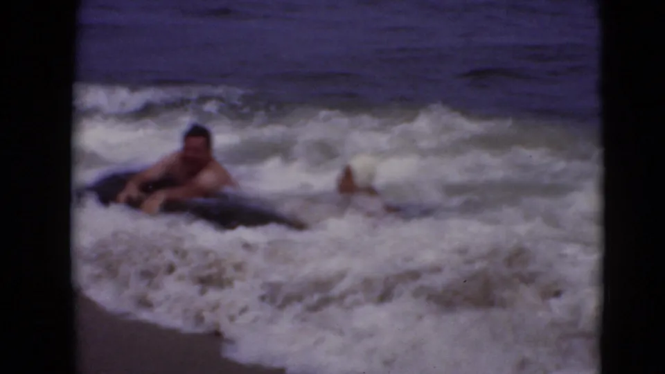 1940s 8mm home-movie frame of two adults partly submerged in foamy shoreline surf, with one white bathing cap visible.