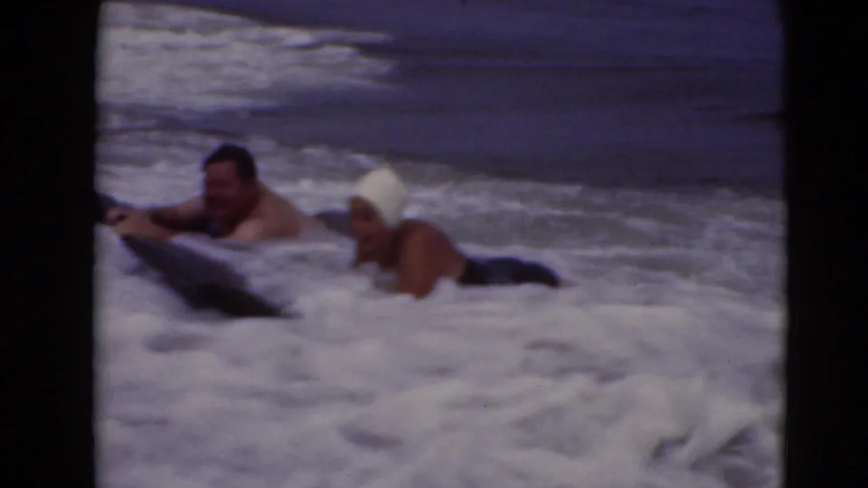 1940s 8mm frame of two adults in rough surf, one with a white bathing cap, drifting beside a black bodyboard.