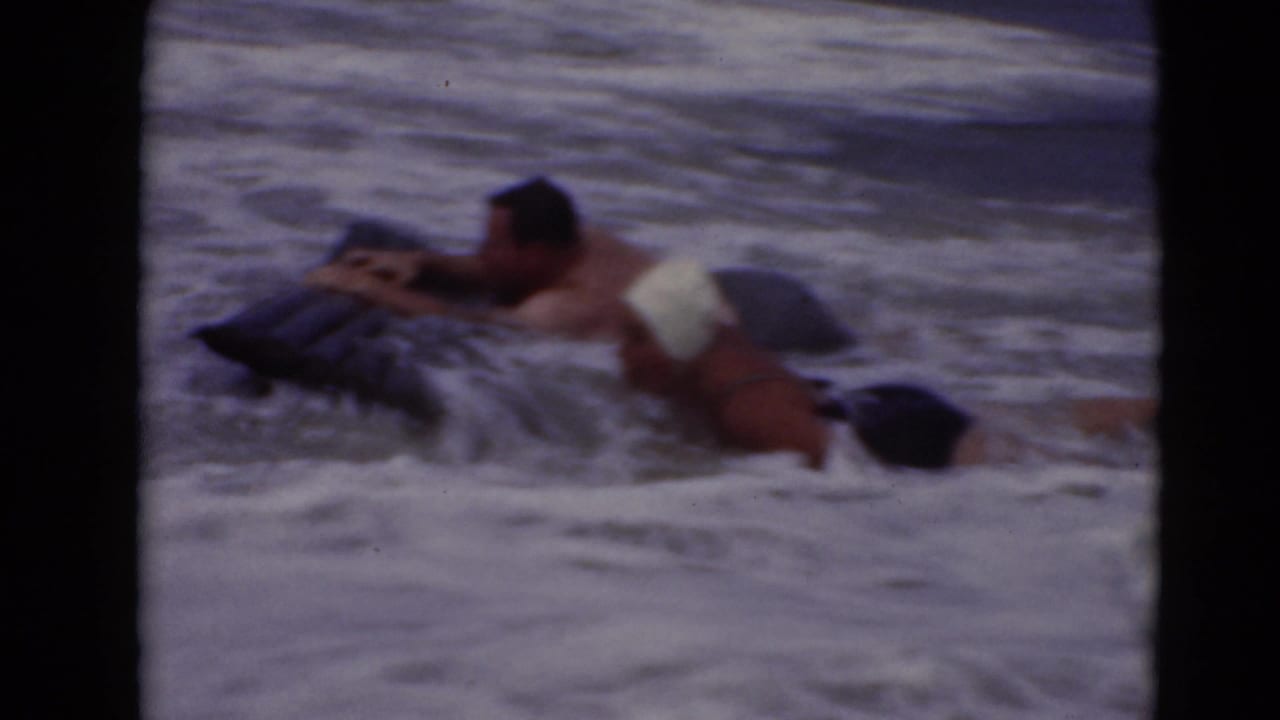 In 1947 home-movie footage, two adults appear in breaking surf with a dark pool float, one wearing a white swim cap.