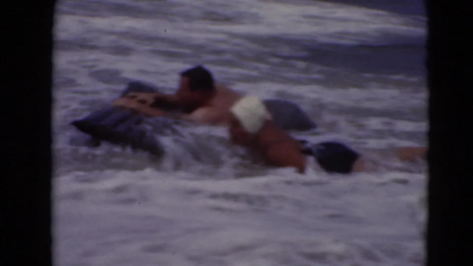 In 1947 home-movie footage, two adults appear in breaking surf with a dark pool float, one wearing a white swim cap.