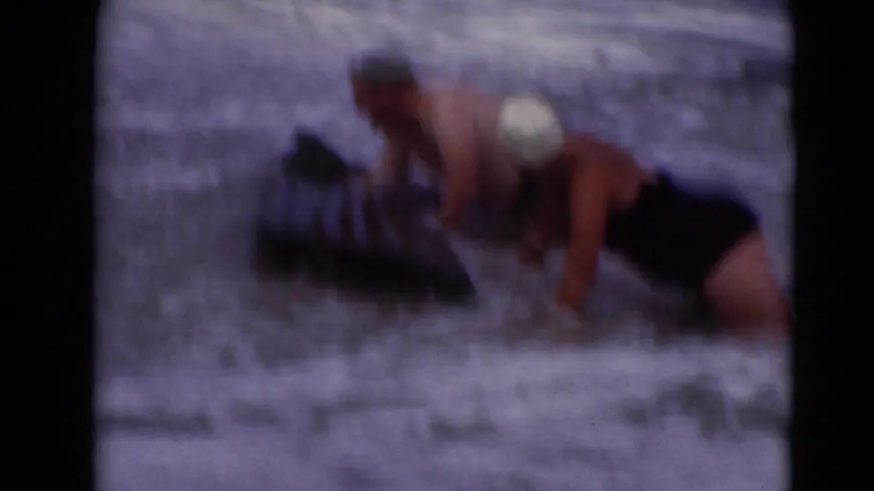 An adult in a white swim cap leans over a dark pool float as another adult is partly visible in the surf.