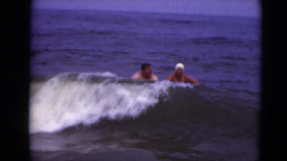 Two adults appear behind a low breaking wave, with the white swim cap and dark float partly visible in soft 8mm color.