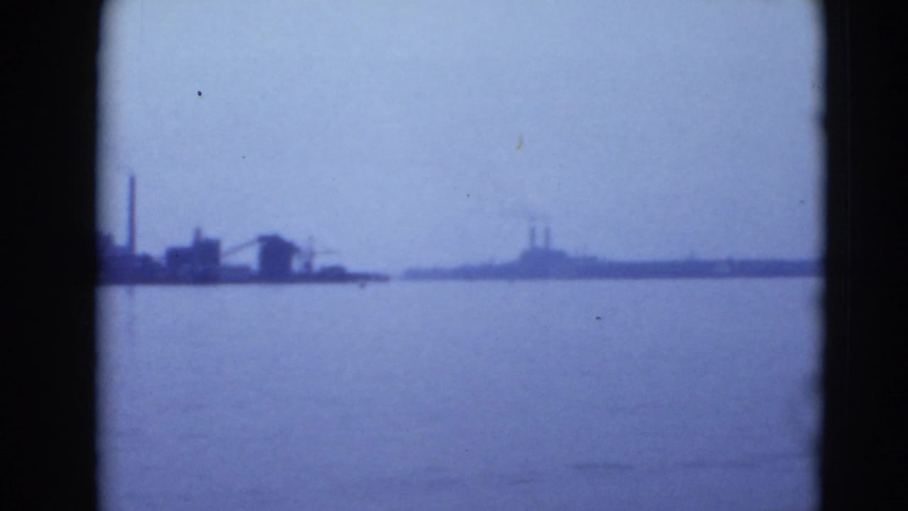 Distant industrial shoreline across calm water under a cloudy sky.