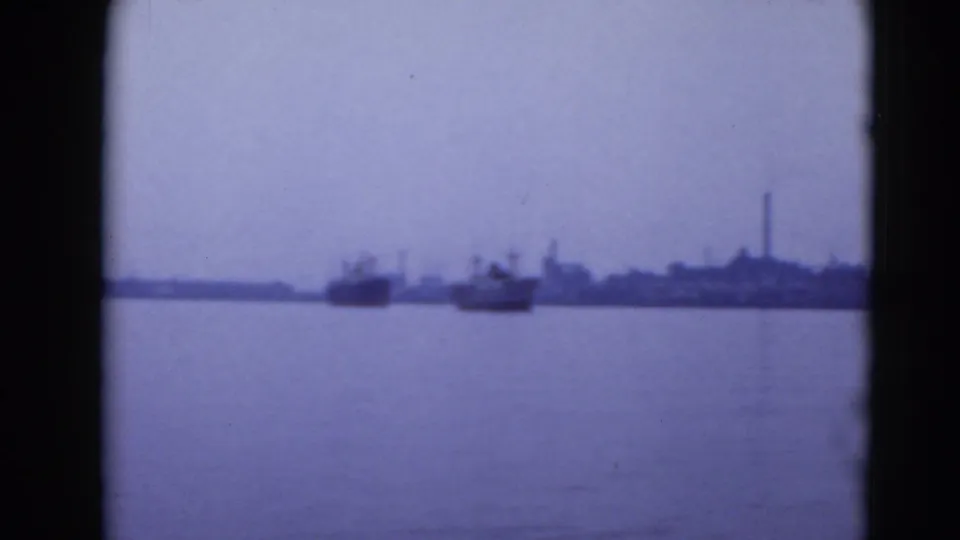 Distant boats on calm water with a low industrial shoreline and tall smokestack in archival 8mm footage.