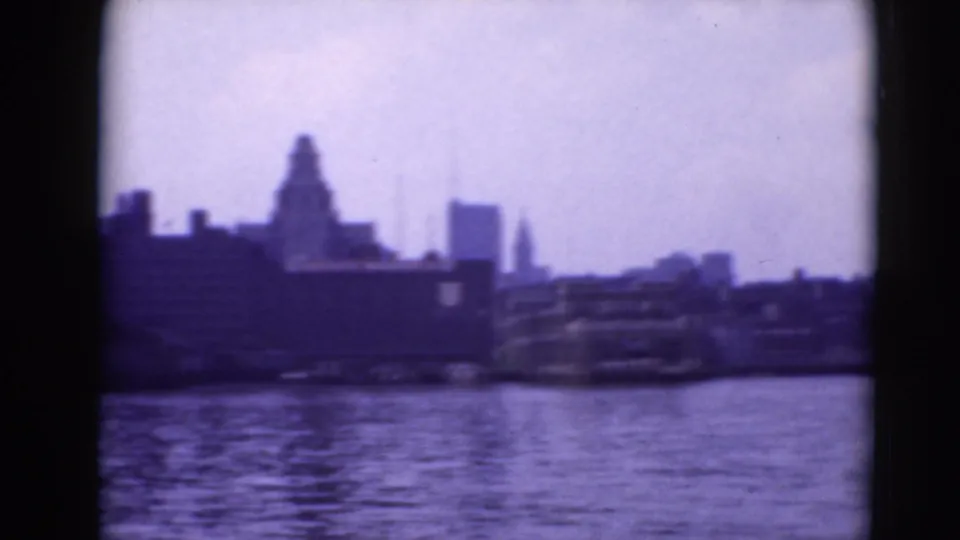 Soft 1940s 8mm view of a waterfront skyline across rippled water, with dark frame borders and a purple cast.