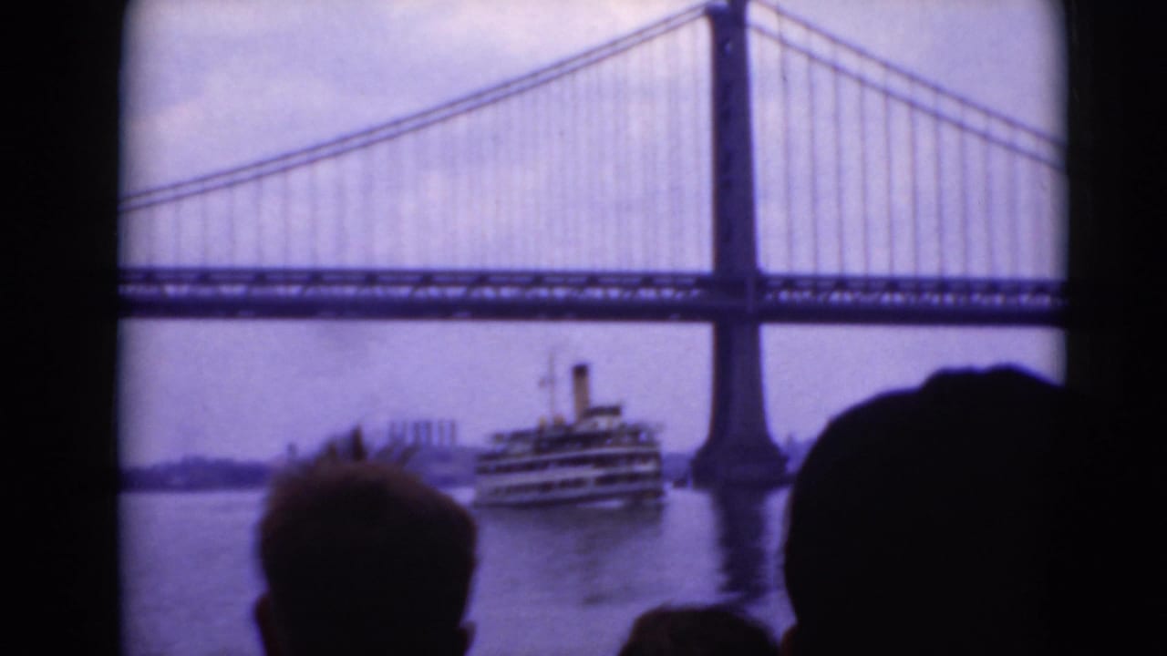 Suspension bridge over a river with a large passenger boat and silhouetted heads in the foreground.
