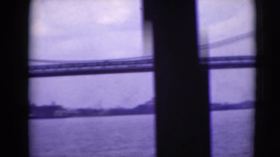 The camera frames the suspension bridge span across the river, partly interrupted by a dark vertical film or vessel edge.