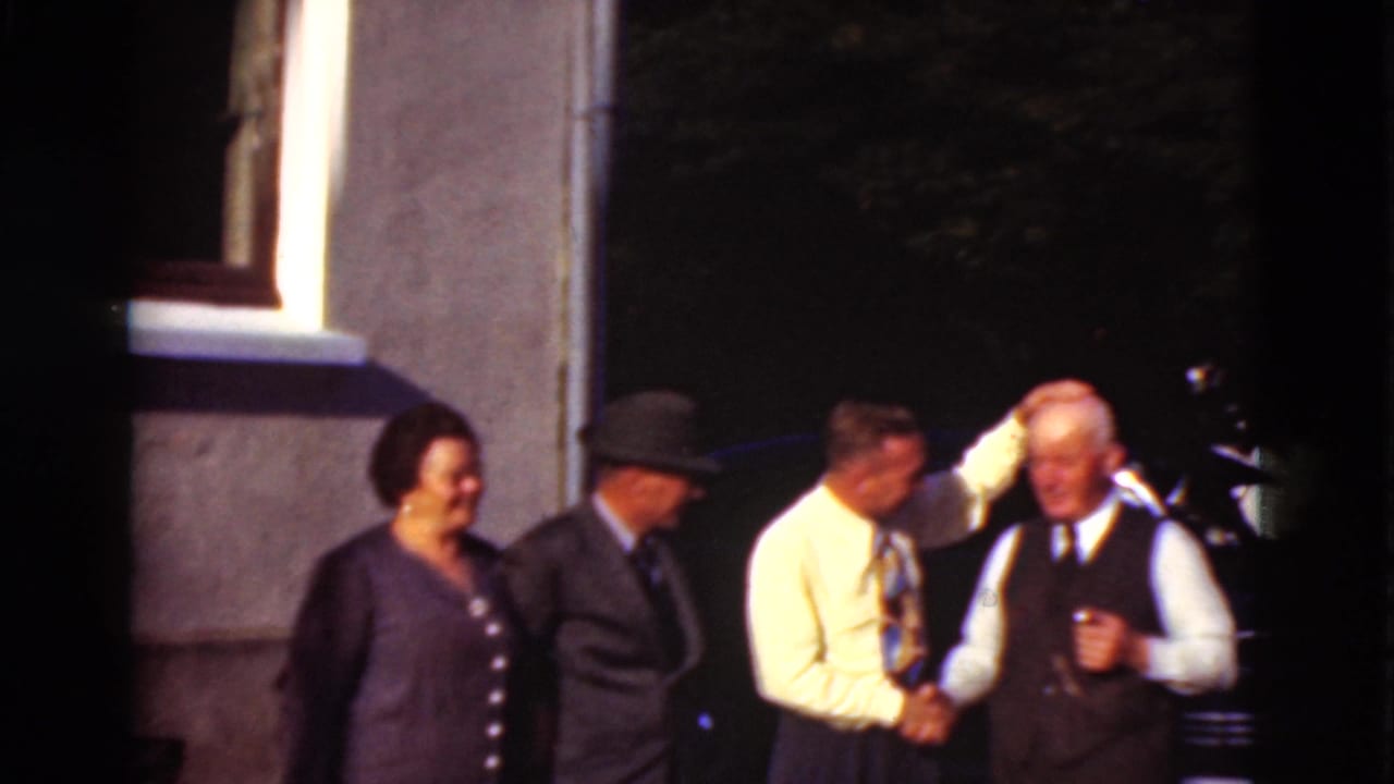Four adults stand outdoors beside a building as two men greet each other with a handshake.
