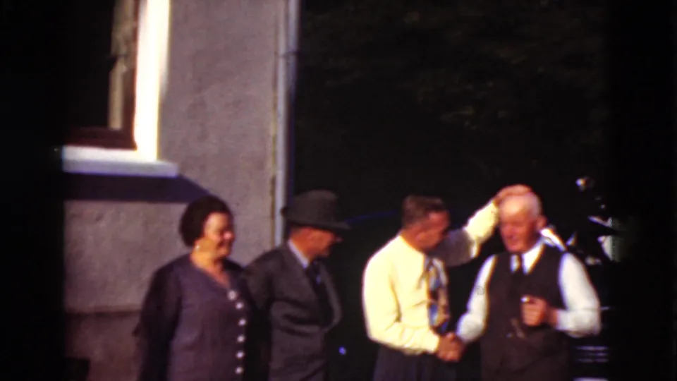 In Denmark 1975 home-movie footage, four adults stand near a building while one man shakes hands with an older man and another reaches toward him.