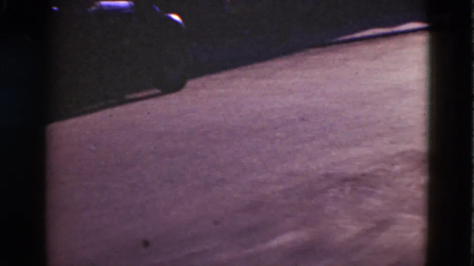 1975 Denmark 8mm frame focused on the road surface, with a shadowed curb and a partial parked car along the upper edge.