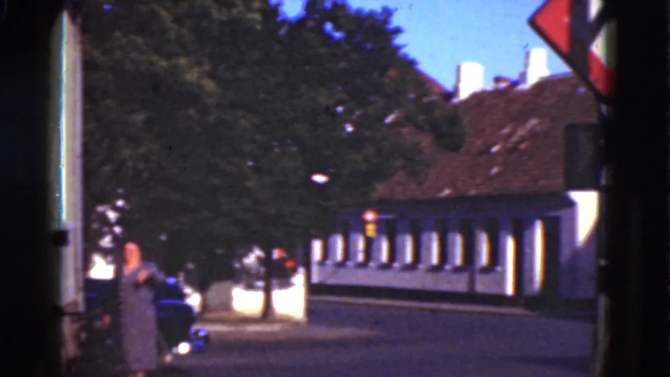 1975 Denmark 8mm frame showing a woman walking and waving near a quiet street, with trees, parked cars, and a tiled-roof building behind her.