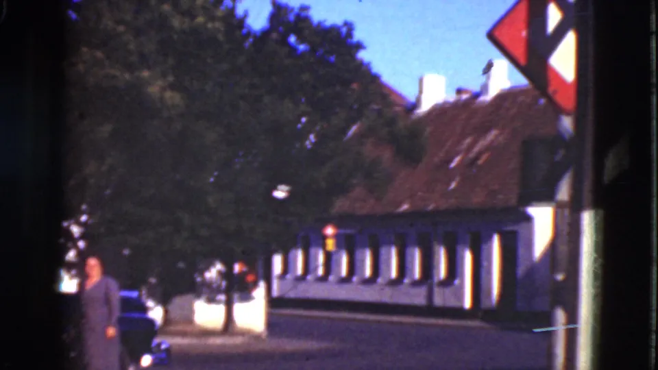 1975 Denmark 8mm frame of a quiet street with white buildings, red-brown roofs, trees, a sidewalk pedestrian, and a partial car.