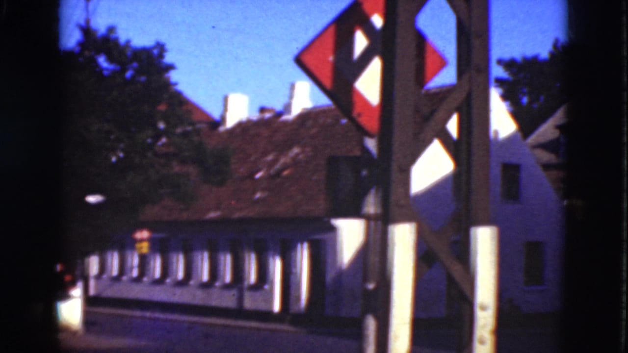White buildings with tiled roofs seen behind a roadside signpost and trees.