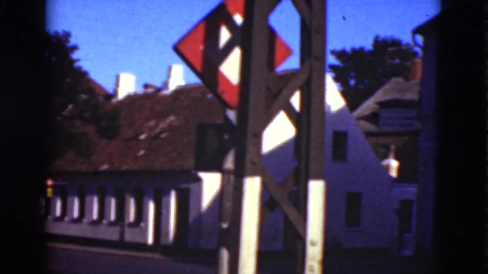 1975 Denmark 8mm frame with a roadside signpost in front of white buildings, tiled roofs, trees, and deep blue shadows.