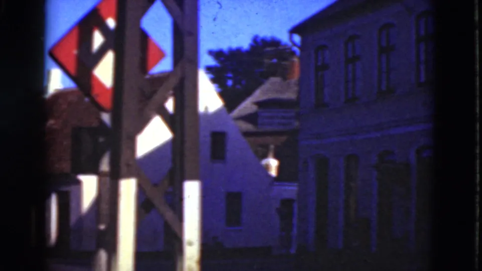1975 Denmark 8mm frame of a red-and-white signpost with white houses and a darker multi-window building at the street corner.