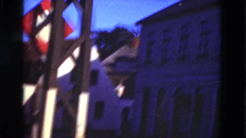 1975 Denmark home-movie frame looking past a wooden structure and red-and-white flag toward pale rural buildings under deep blue sky.