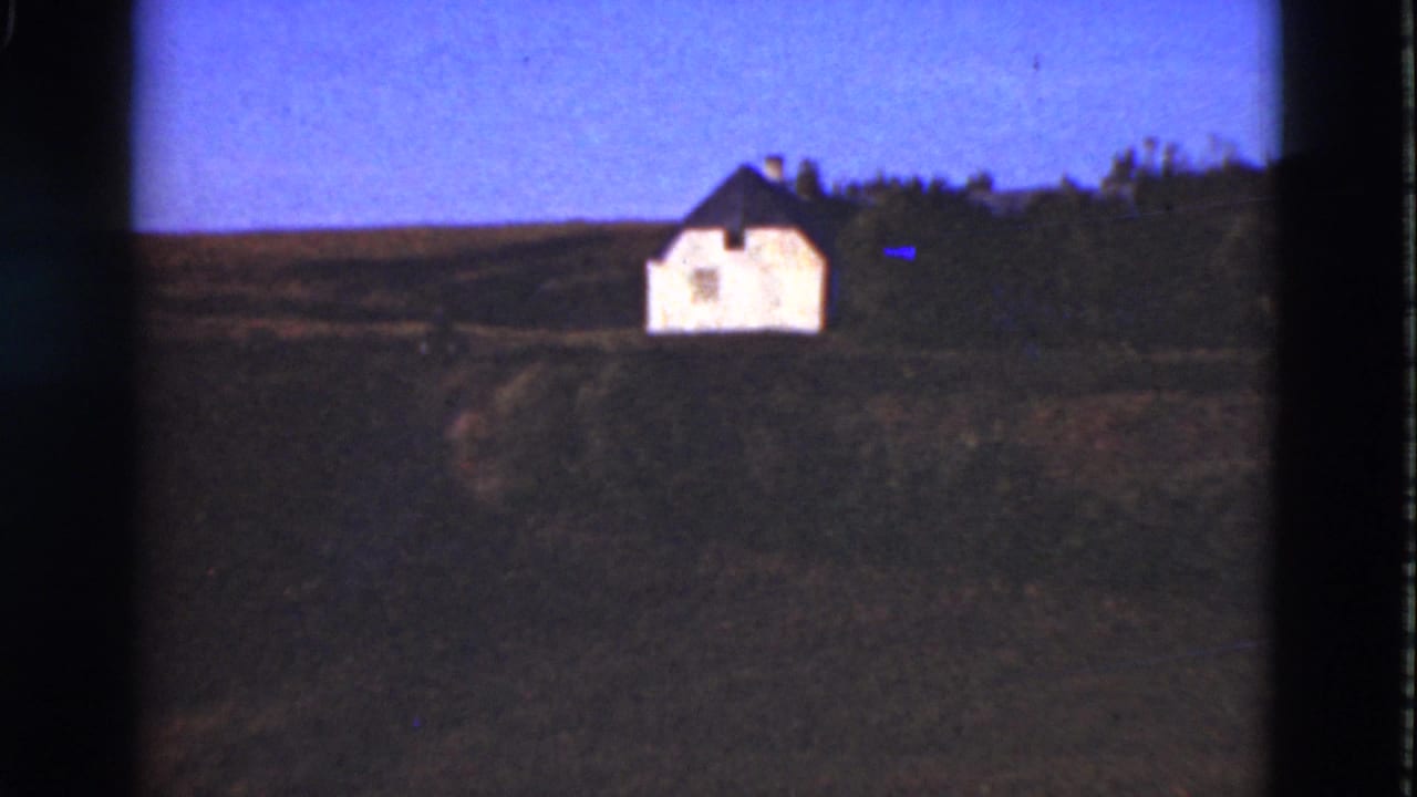 Distant white house on a grassy hillside with fields and trees behind it.